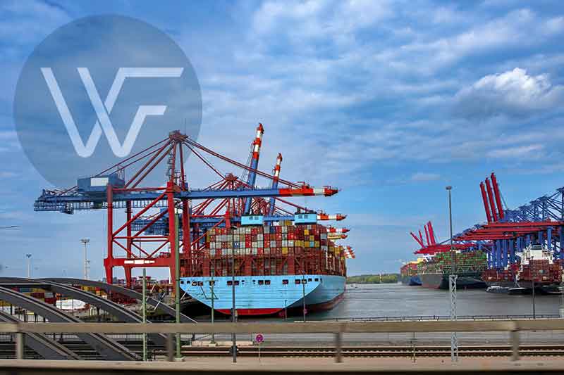 A large container ship docked at a port with multiple cranes in the background.