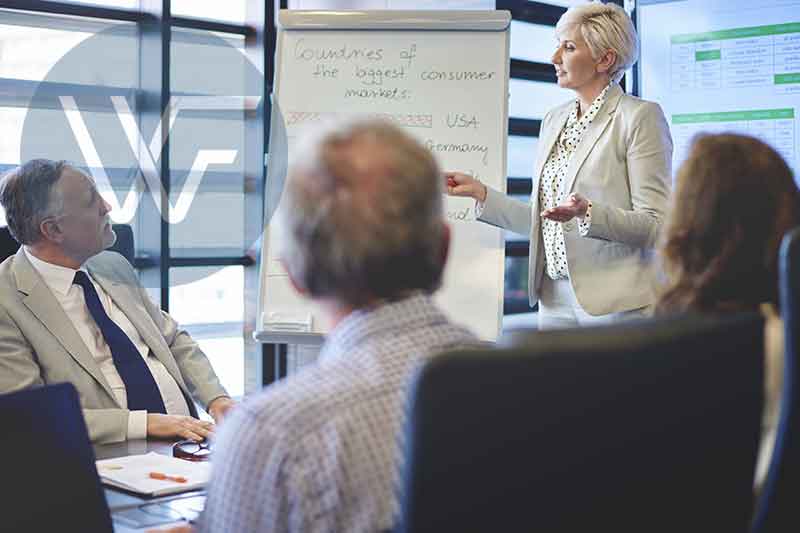 A woman presenting in a conference room with a whiteboard and audience members seated at a table.