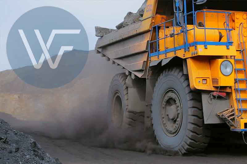 A large yellow mining truck transporting materials on a dirt road, kicking up dust.