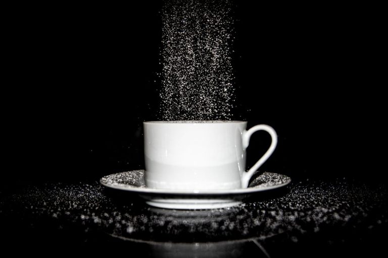 Dynamic black and white image of sugar pouring into a white cup on a saucer against a black background.