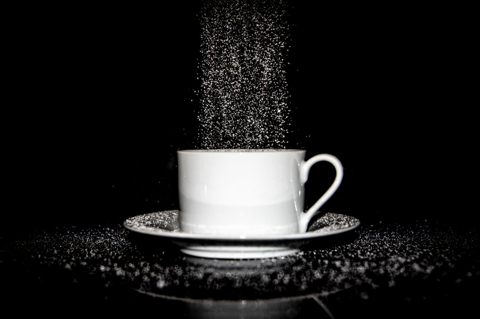 Dynamic black and white image of sugar pouring into a white cup on a saucer against a black background.