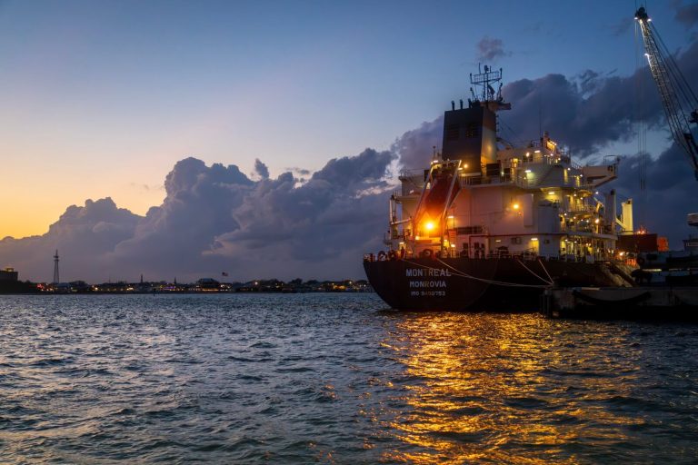 A cargo ship moored in a harbor during sunset, reflecting lights on the water.