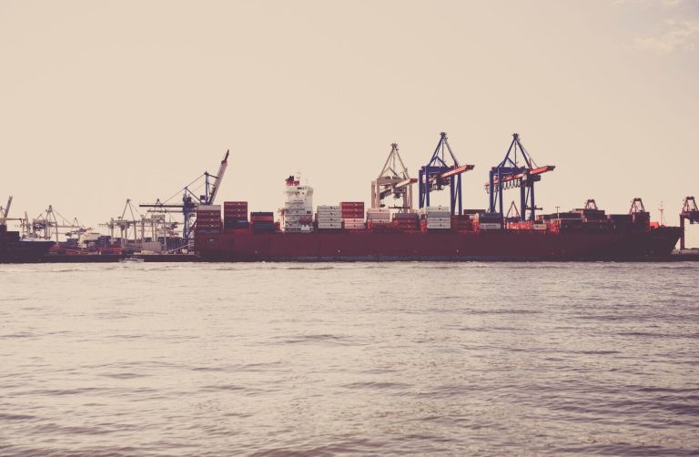 A cargo ship loaded with containers docked at a seaport under clear skies.