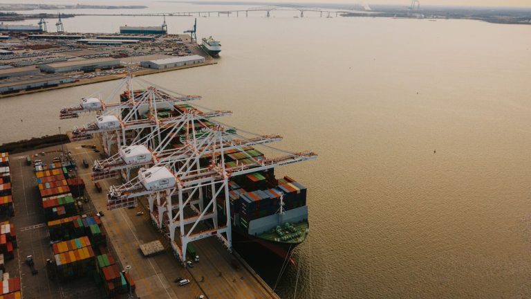 Drone view of cargo containers loaded on long ship on river of pier with lifting cranes