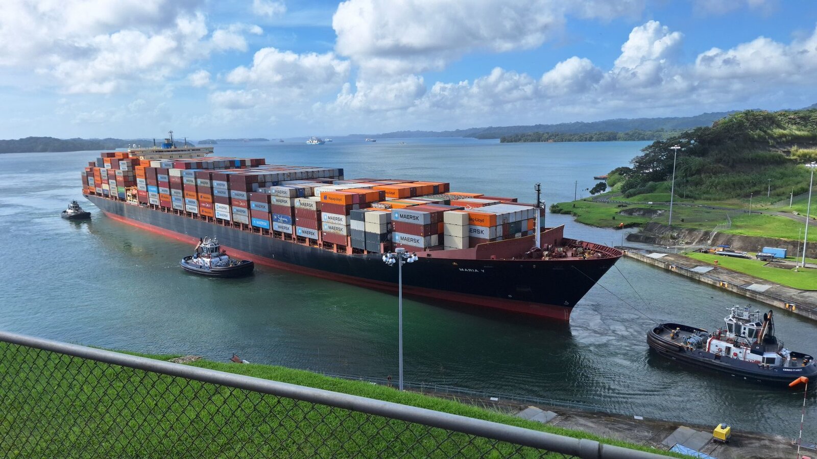 Large container ship maneuvered by tugboats through the Panama Canal under a clear sky.