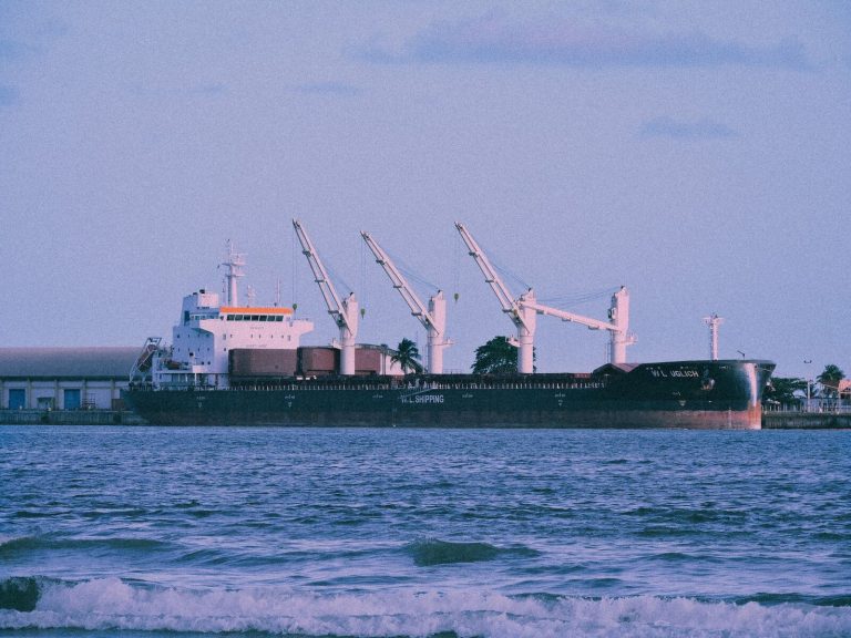 Large cargo ship docked at a port, cranes visible, with calm sea foreground.