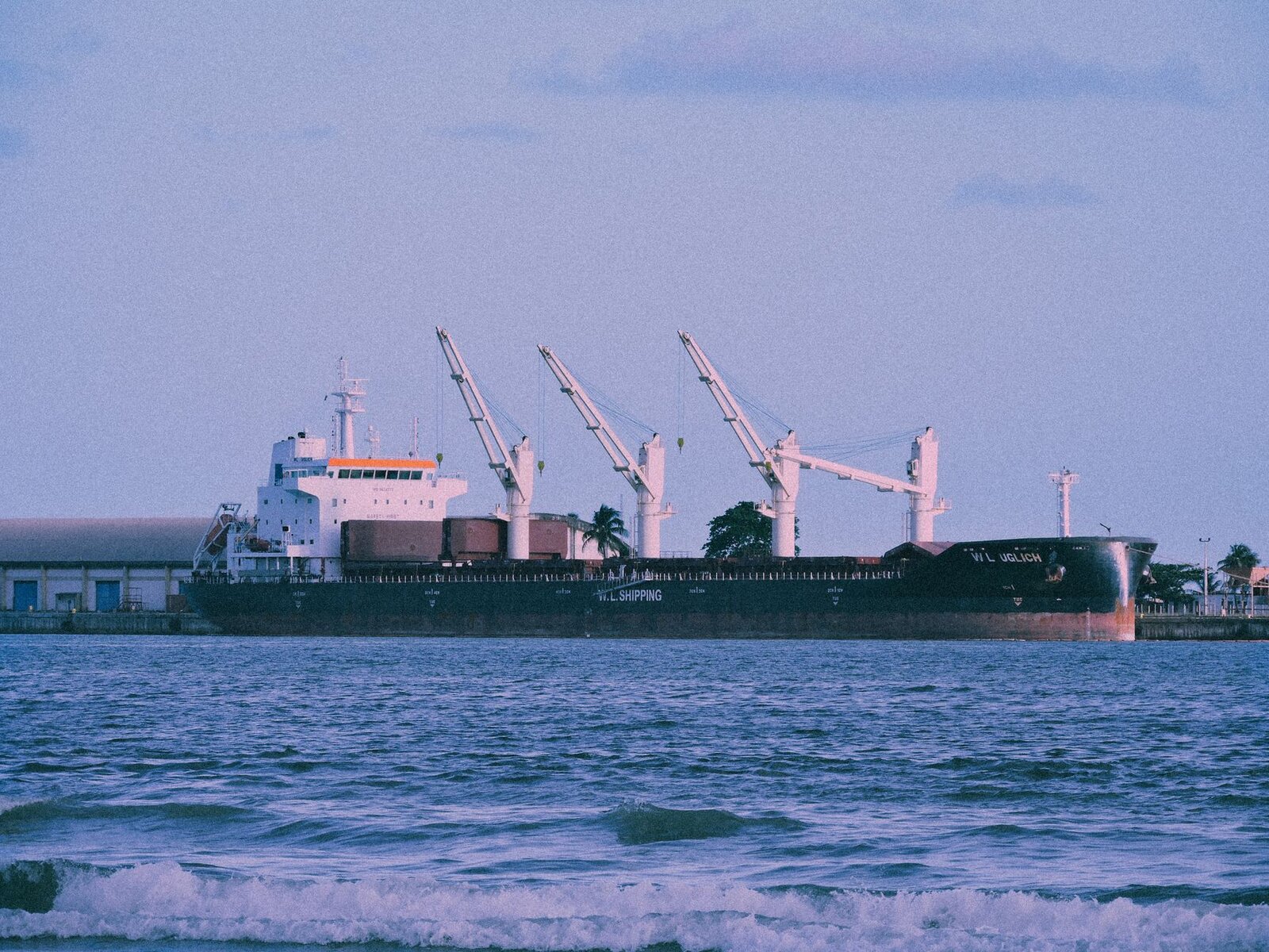 Large cargo ship docked at a port, cranes visible, with calm sea foreground.