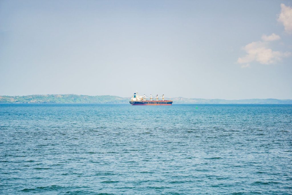 A cargo ship sails on a calm sea against distant green hills under a clear blue sky.