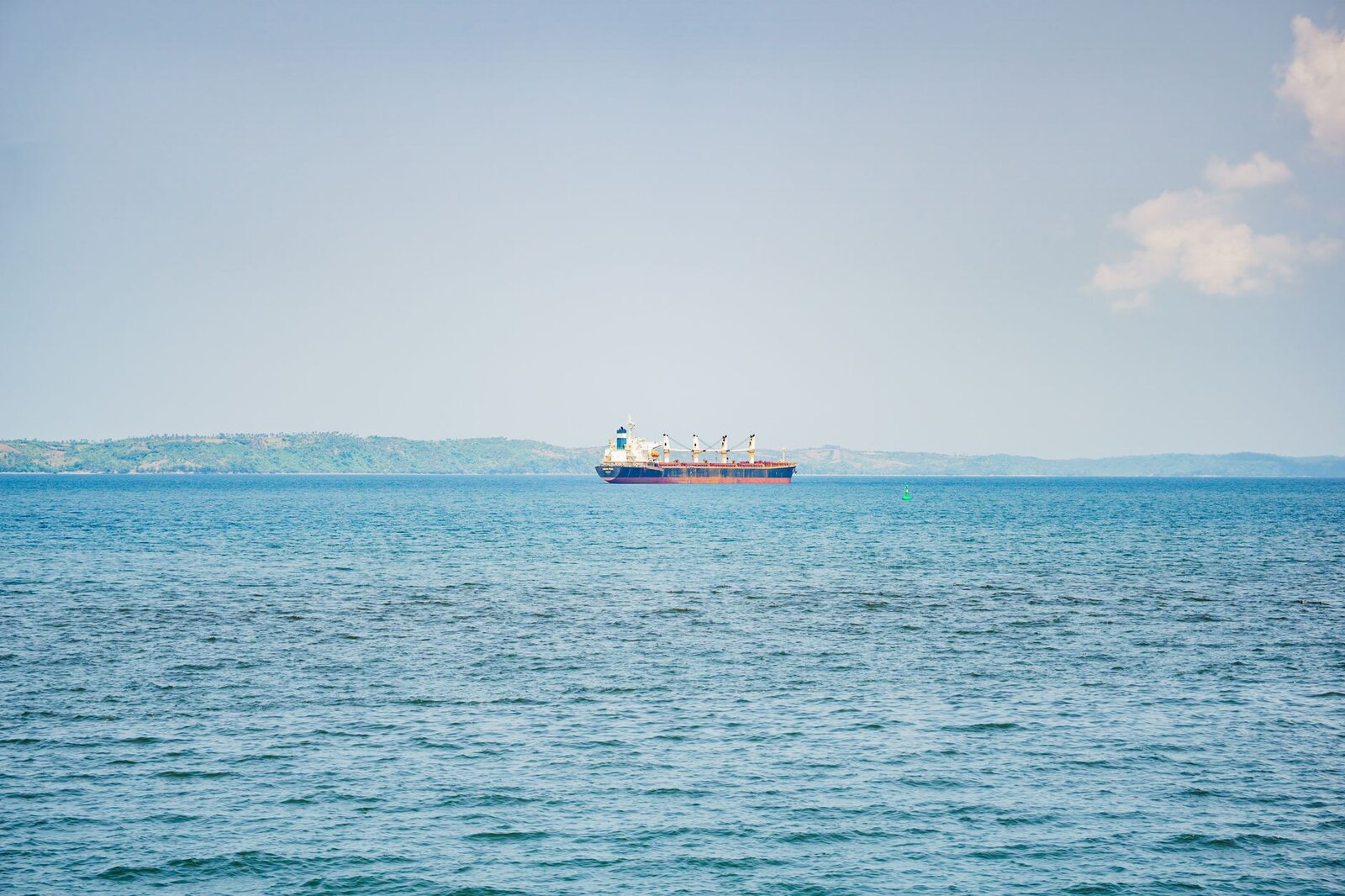 A cargo ship sails on a calm sea against distant green hills under a clear blue sky.