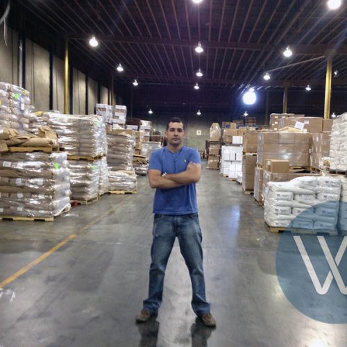 A warehouse worker standing with arms crossed in a storage area filled with stacked boxes and bags.