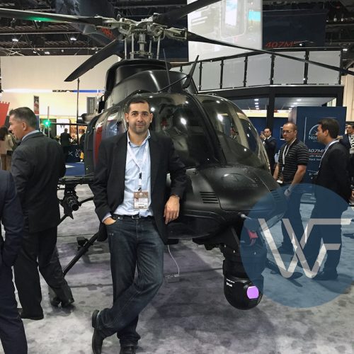 Individual standing beside a black helicopter at an exhibition event with attendees in the background.