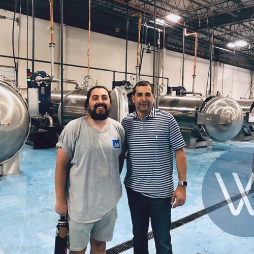 Two men posing for a photo in an industrial facility with large stainless steel equipment in the background.