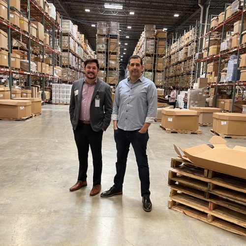 Two men standing in a warehouse aisle surrounded by stacked boxes and pallets.